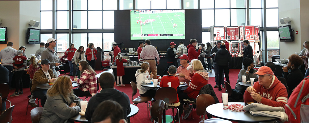 People seated around large round tables in front of a large screen tv showing the football game